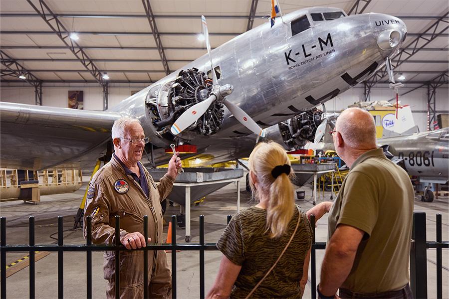 Man en vrouw krijgen een rondleiding in een museum over vliegtuigen en kijken naar een oud model van een KLM-vliegtuig