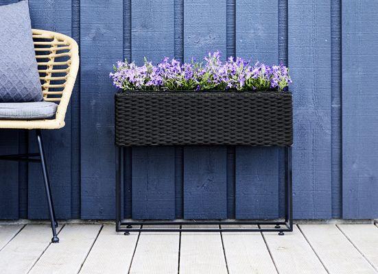 Zwarte rotan plantenbak met bloemen erin met een stoel ernaast voor een muur