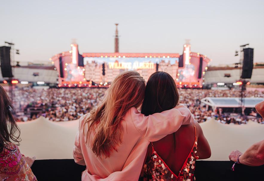 Twee meiden met een arm om elkaar heen van bovenaf uitkijkend op een groot podium met groot publiek