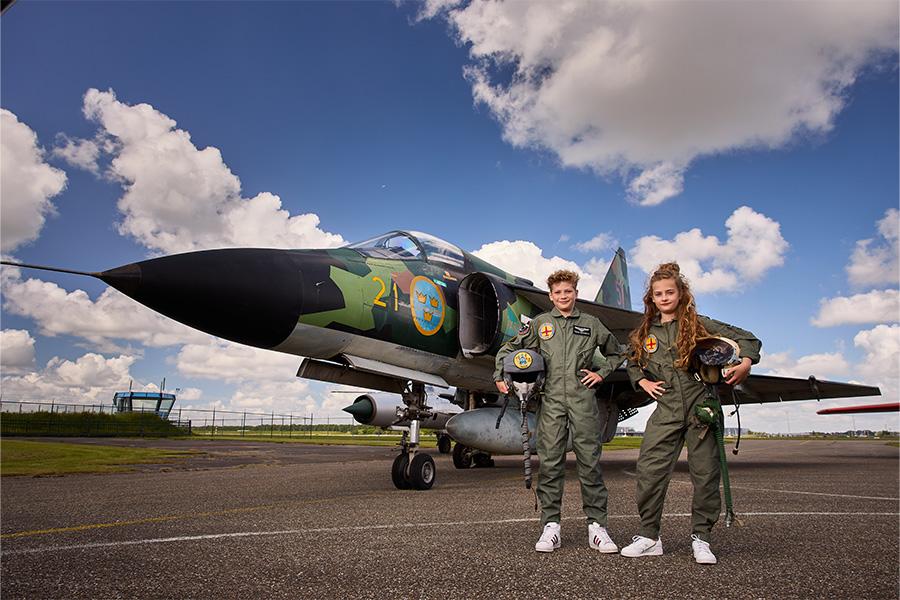 Twee kinderen in overall met helm onder hun arm voor een vliegtuig met camouflageprint