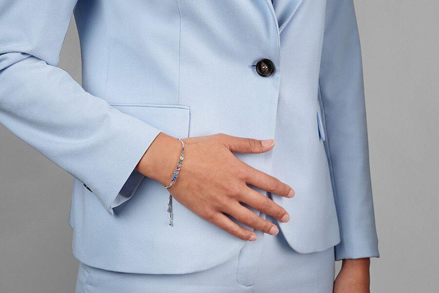 Hand met zilverkleurige arrmband met meerkleurige steentjes die wordt gedragen door een persoon in een lichtblauw pak